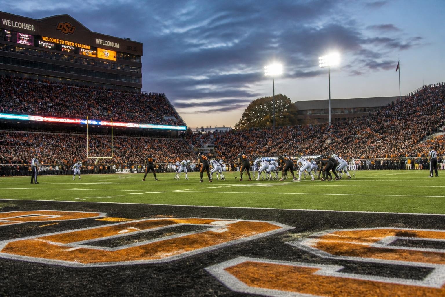 College football stadium packed with fans during evening matchup with field-level view of players and bright stadium lighting